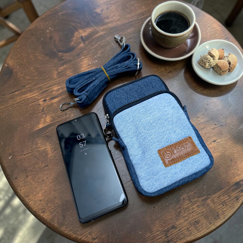 Bidora two-tone blue denim crossbody mobile phone pouch with detachable strap on a rustic wooden table with coffee.
