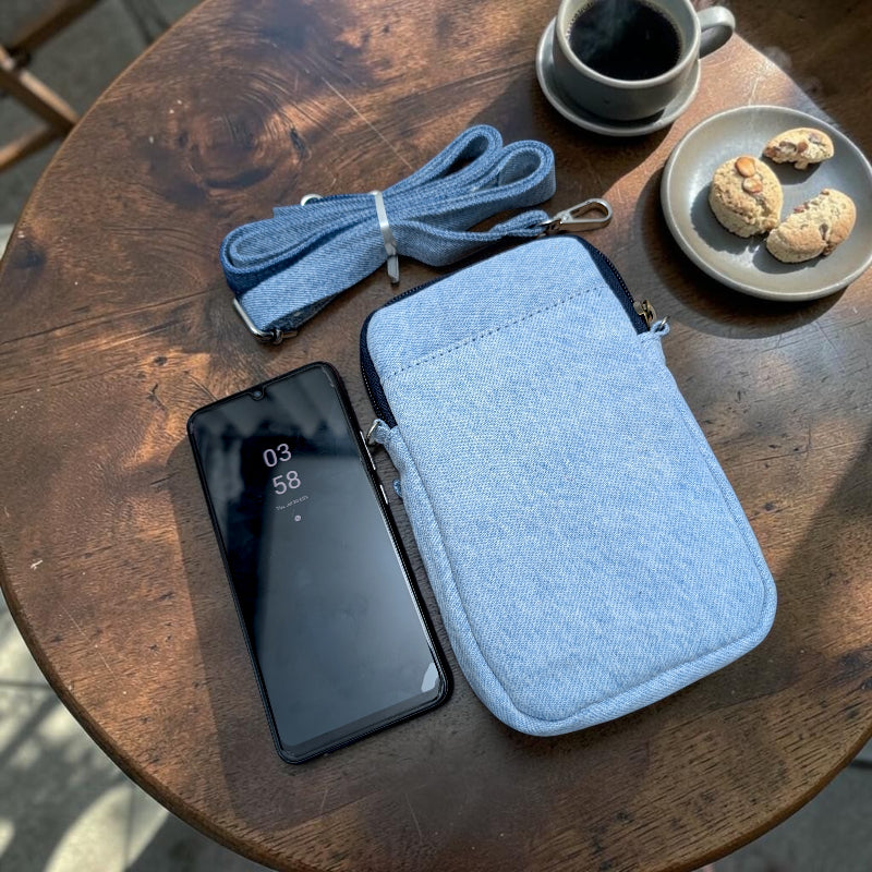 Bidora light blue denim crossbody phone pouch with detachable strap and smartphone on a rustic wooden table