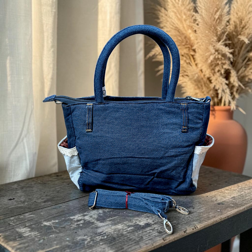 Bidora blue denim handbag with top handles, side pockets with lace trim, and a detachable shoulder strap on a table.