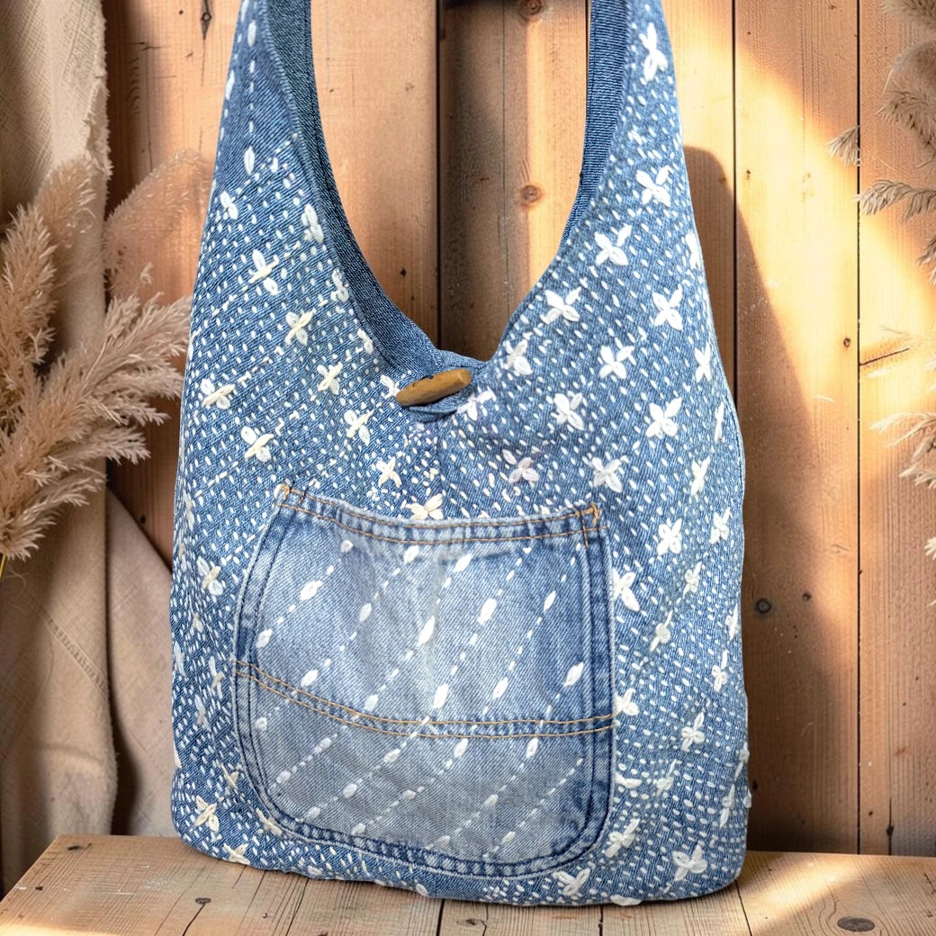 Bidora denim hobo bag with white hand-stitched embroidery, exterior pocket, and rustic wooden toggle closure.