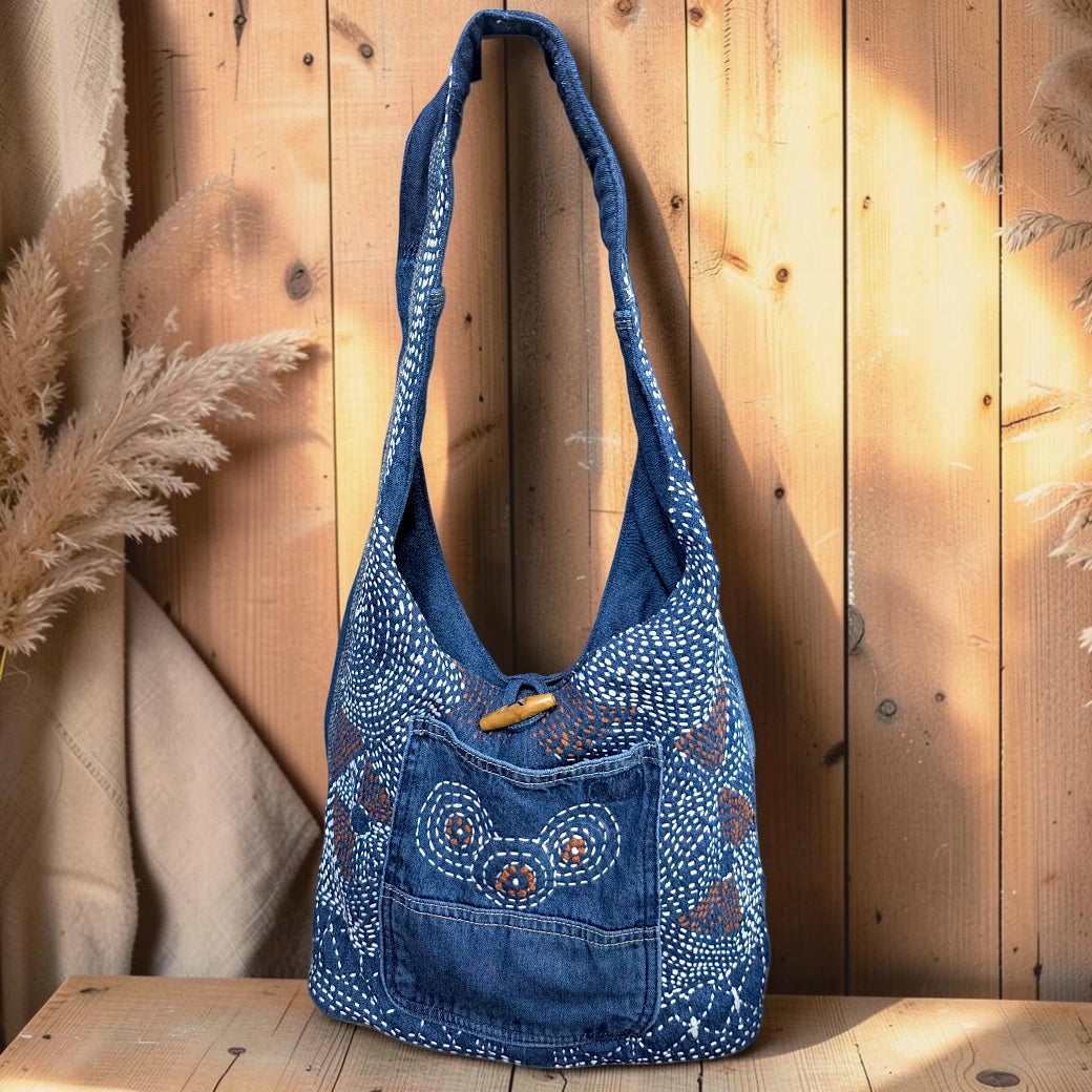 Bidora indigo denim hobo shoulder bag with intricate white hand-stitched embroidery and a rustic wooden toggle closure.