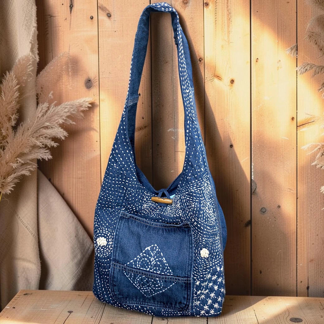 Bidora indigo denim hobo shoulder bag with white hand-stitched embroidery, wooden toggle closure, and front pocket.