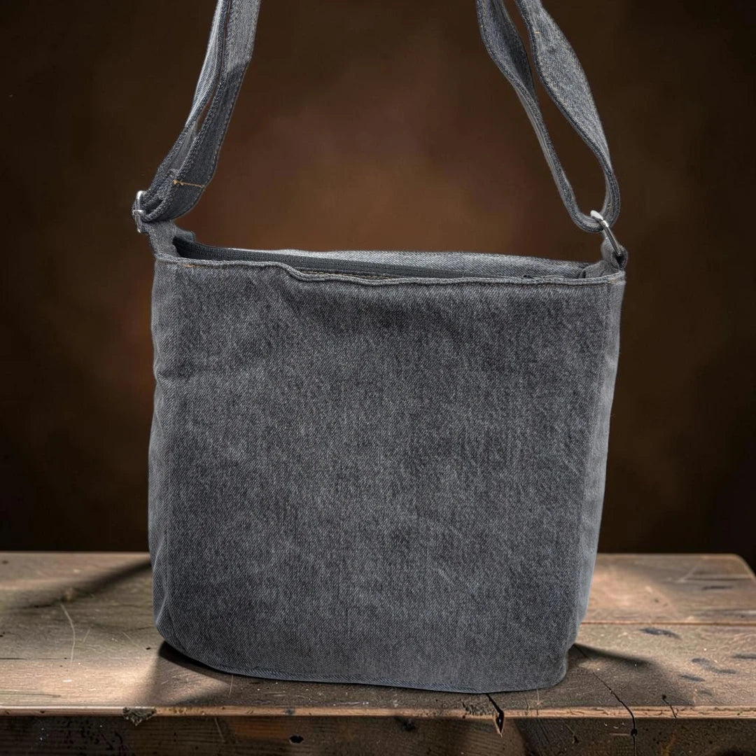 Bidora grey upcycled denim crossbody shoulder bag with an adjustable strap sitting on a rustic wooden surface.
