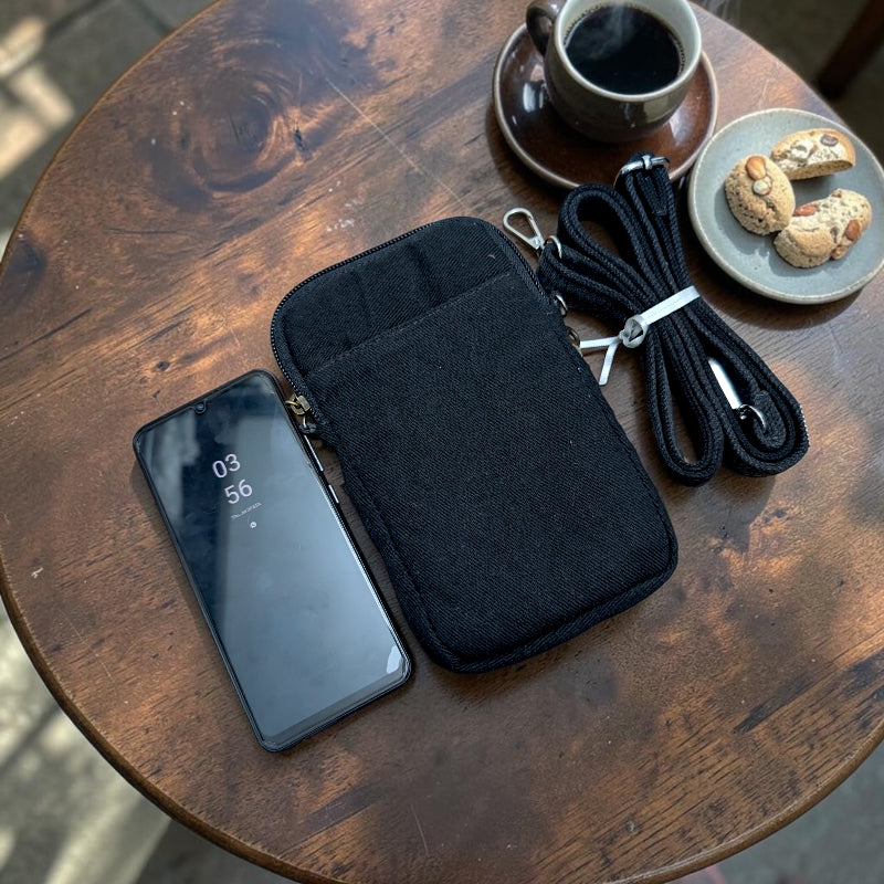 Bidora black fabric crossbody phone pouch with adjustable strap, shown on a wooden table next to a smartphone.