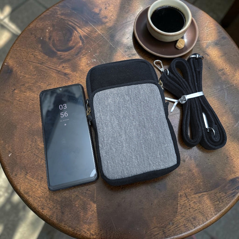 Bidora two-tone black and grey fabric crossbody phone pouch with adjustable strap on a wooden table next to a phone.