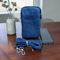 Bidora denim crossbody phone pouch with detachable strap and zippered compartments on a wooden table next to a phone.