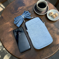 Bidora light blue denim crossbody mobile phone pouch with an adjustable strap resting on a wooden cafe table.