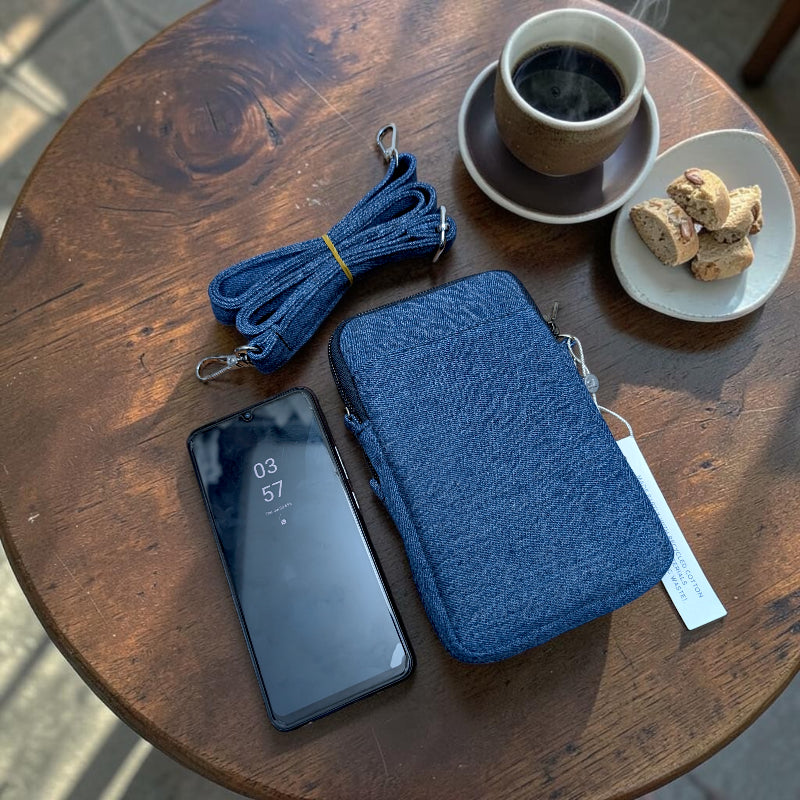Bidora denim crossbody mobile phone pouch with detachable strap, shown on a wooden table with a smartphone and coffee.