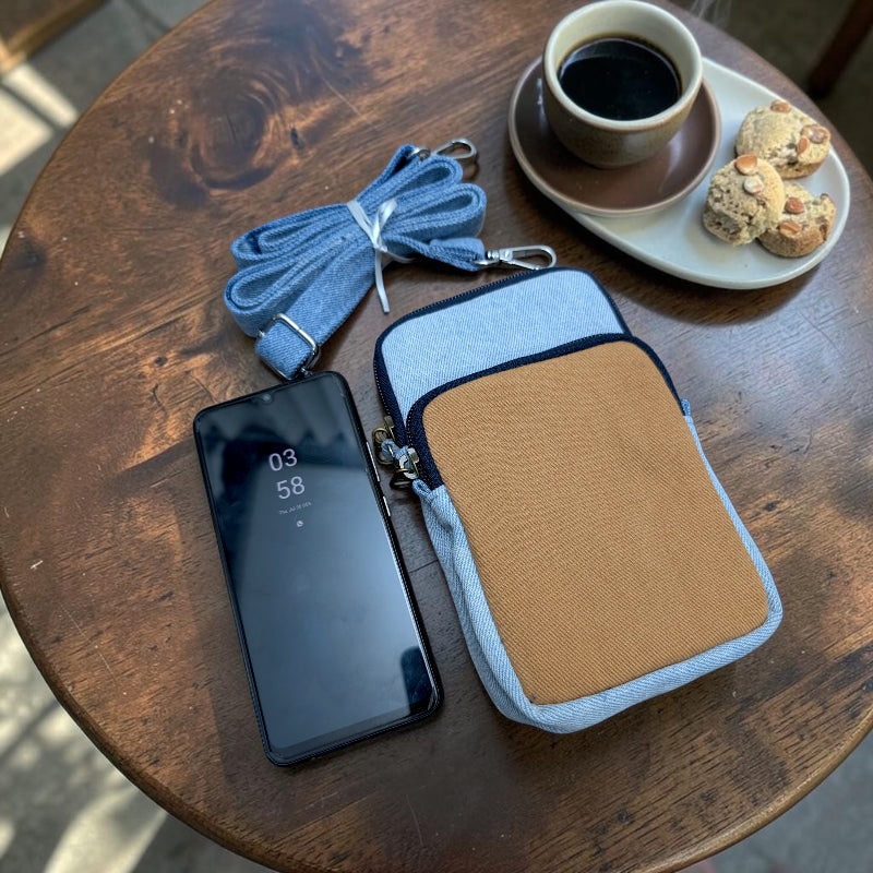 Bidora casual denim crossbody phone pouch with tan pocket, adjustable strap, and smartphone on a wooden table.