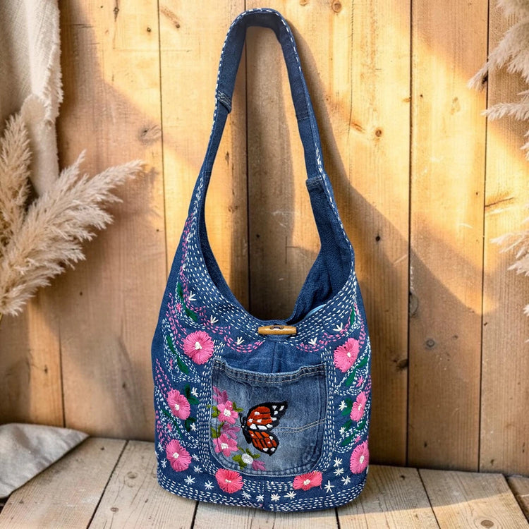 Bidora denim hobo shoulder bag with floral embroidery, butterfly motif, and wooden toggle closure on a wooden background