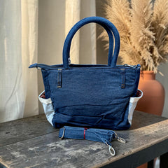 Bidora blue denim handbag with top handles, side pockets with lace trim, and a detachable shoulder strap on a table.