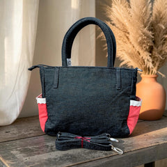 Bidora dark denim handbag with red side panels, white trim, top handles, and a detachable shoulder strap on a table.