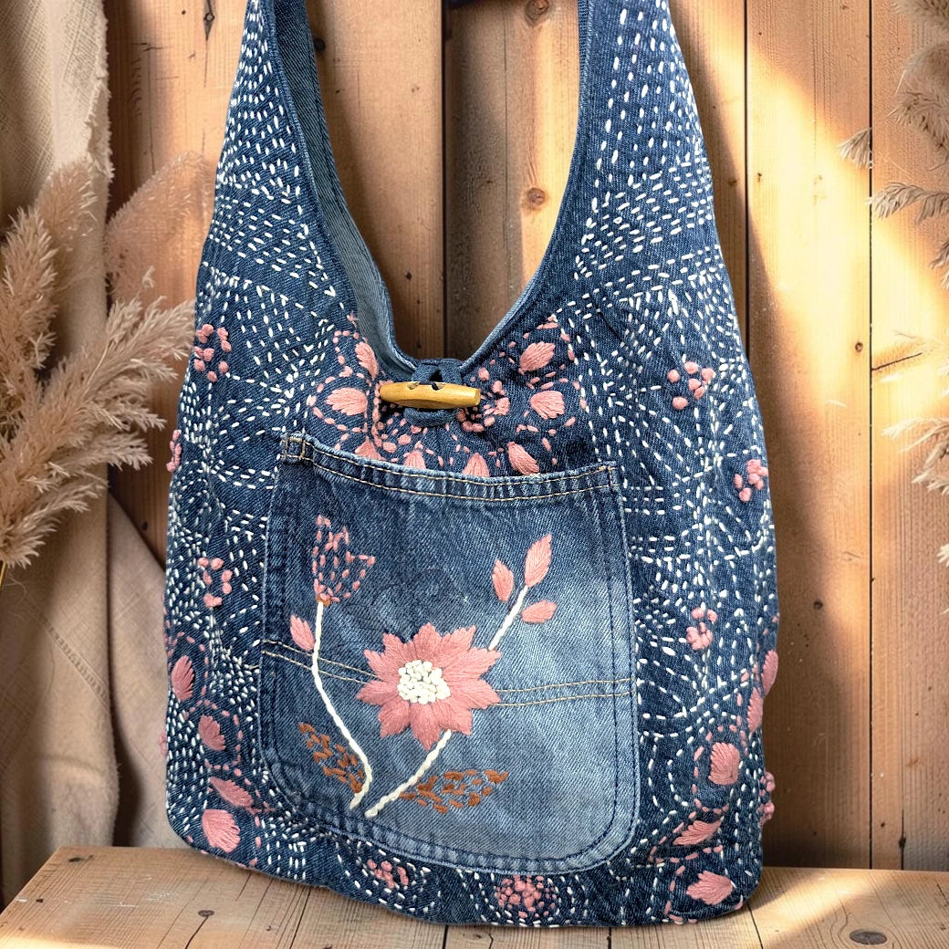 Bidora indigo denim hobo bag featuring pink floral embroidery, white stitching, and a wooden toggle closure.