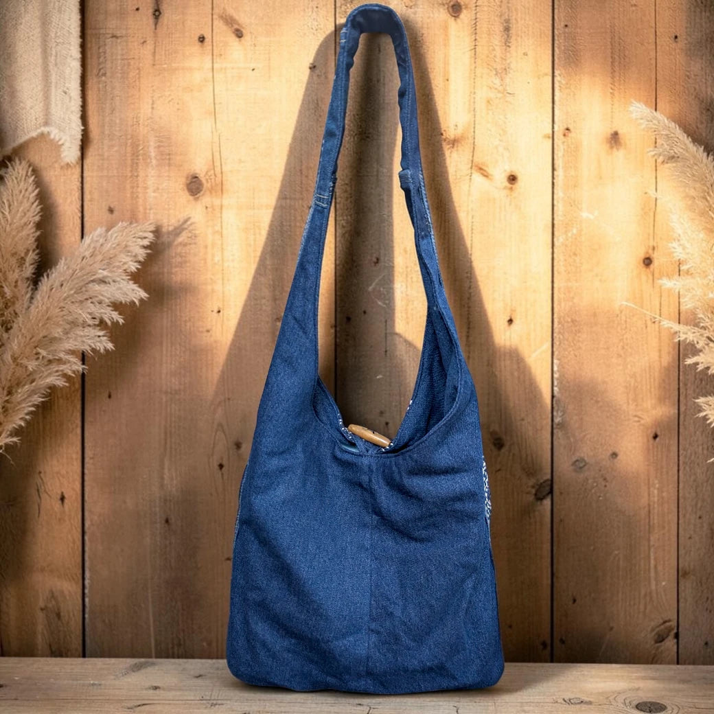 Bidora indigo denim hobo shoulder bag with a wide strap and rustic wooden toggle closure against a wooden background.