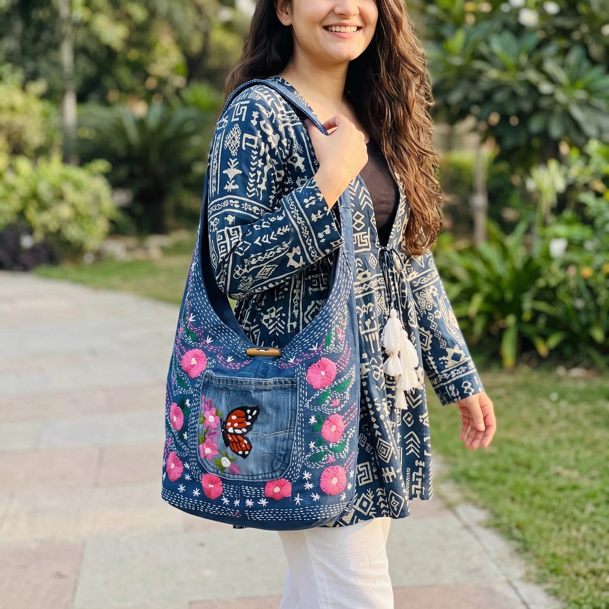 Bidora denim hobo shoulder bag with pink floral embroidery and butterfly motif, worn by a woman in a park.