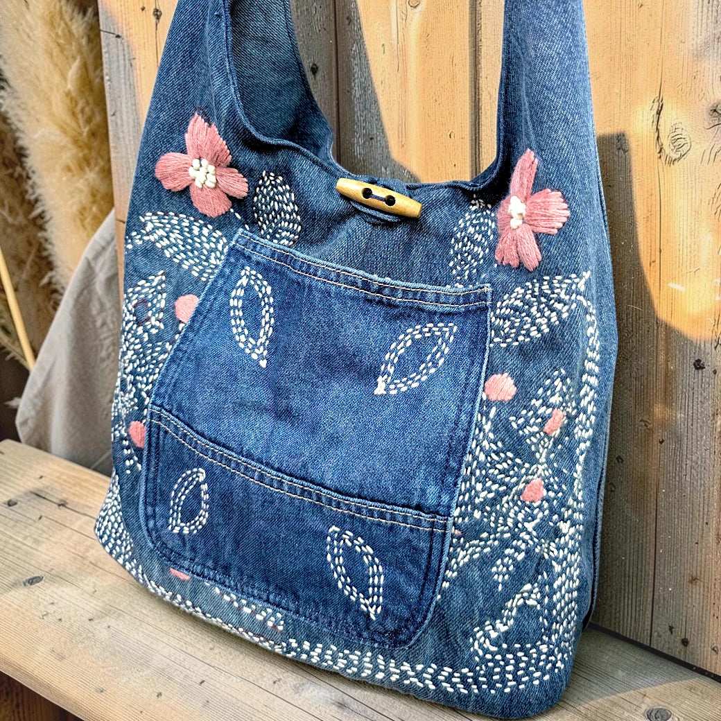 Bidora blue denim hobo bag with pink and white floral embroidery, front pocket, and a rustic wooden toggle closure.