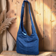 Bidora indigo denim hobo shoulder bag with white hand-stitched embroidery and a wide strap against a wooden background