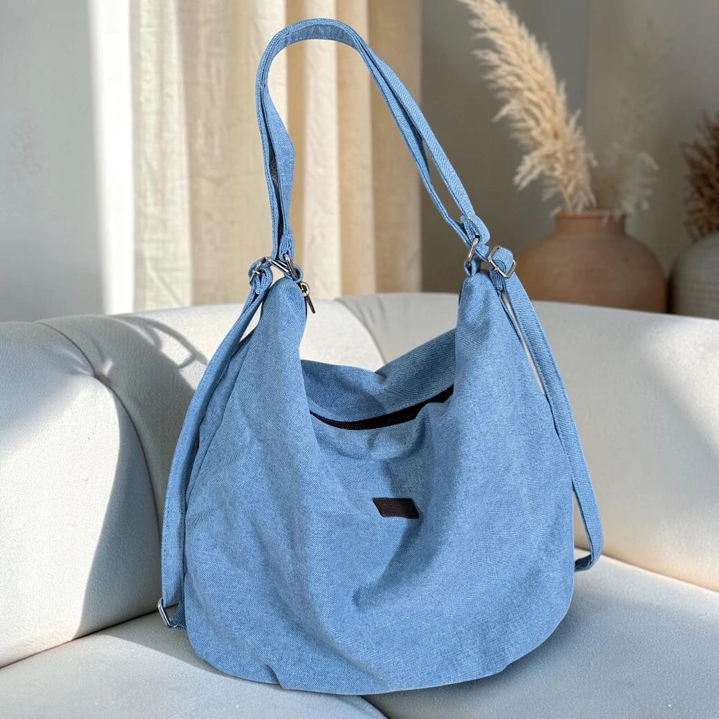 Bidora light blue denim hobo bag with an adjustable strap and secure zipper closure resting on a white sofa.