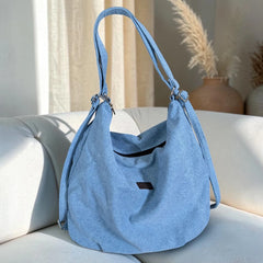 Bidora light blue denim hobo bag with an adjustable strap and secure zipper closure resting on a white sofa.