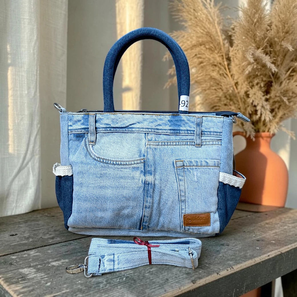 Bidora upcycled light-wash denim tote handbag with dark blue handles, exterior pockets, and detachable shoulder strap.